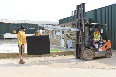 Two of our warehouse staff quality checking a large paving slab. One is operating a forklift holding the slab suspended on a forklift grab. the other staff member is stood next to the slab and is inspecting it for quality assurance.