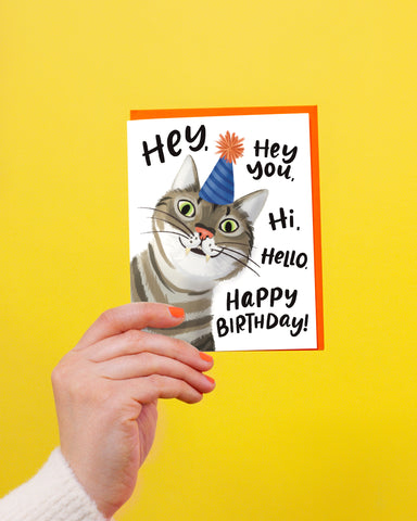 hand holding a Birthday card with an orange envelope. Birthday card has an illustration of a cat in a birthday party hat that says hey, hey you, hi, hello, happy bithday!