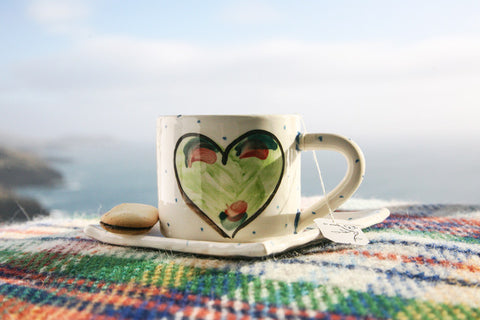 A handmade ceramics pottery mug sits atop a small square ceramics plate. Each has a green heart hand painted on it by Charlie Mahon Ceramics Pottery. Made in Ireland. The duo dit on a plaid blanket with the Irish countryside in the background.