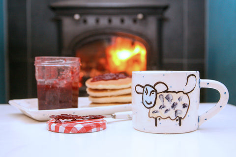 A handmade Irish pottery mug resting on a table. It is hand painted with a smiling sheep. Behind the mug there is a jar of red jam and a stack of pancakes. A roaring fireplace is in the far background. The mug is made in Ireland by Charlie Mahon Ceramics Pottery.