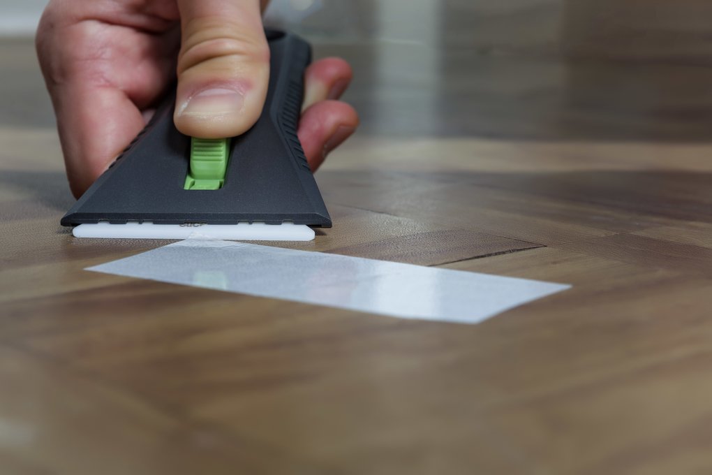 A Slice 10593 Auto-Retractable Ceramic Scraper being used to scrape adhered tape off a wooden surface.