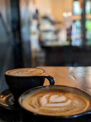 two cups of coffee in black mugs in front of a blurred background of a coffee shop interior