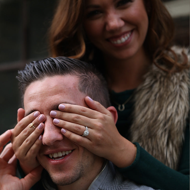 A woman covering another mans eyes wearing a halo ring on her left hand.