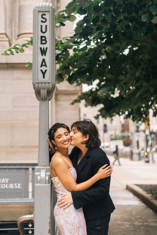 A woman in a suit kissing a woman in a dress on the cheek under a sign for the subway.