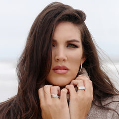 A woman with brown hair wearing a pair of Steven Singer Jewelers Harmonious Huggie Diamond Hoop earrings, and several gold and diamond rings.