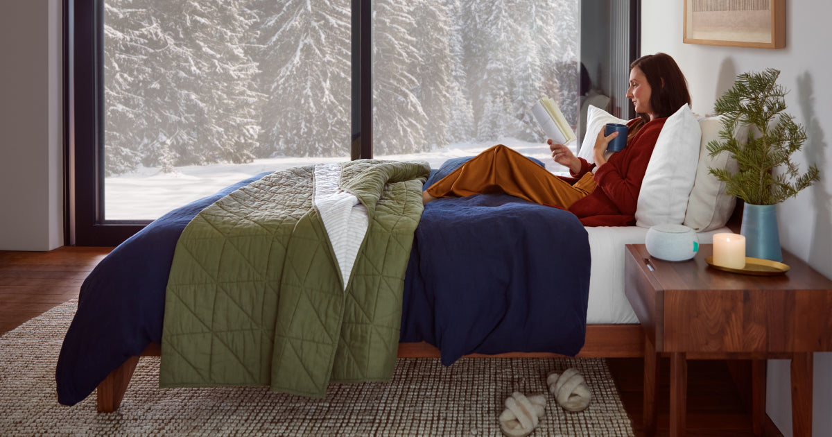 Woman laying in comfy Tuft & Needle bed reading with snow covered trees outside of her window.