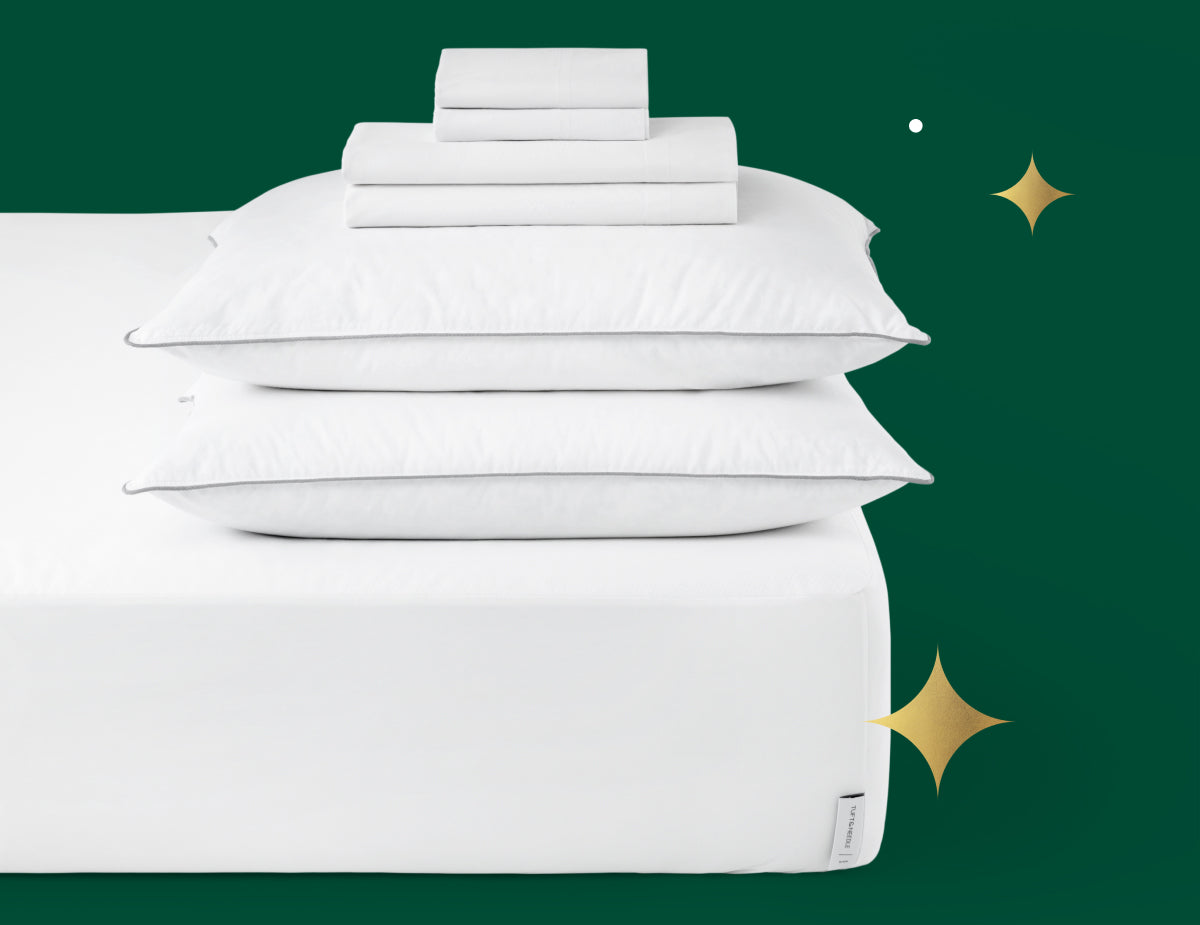 Stacked on top of the Tuft & Needle Original mattress is 2 down alternative pillows, folded sheets and folded pillowcases. Picture has a green background with gold stars.