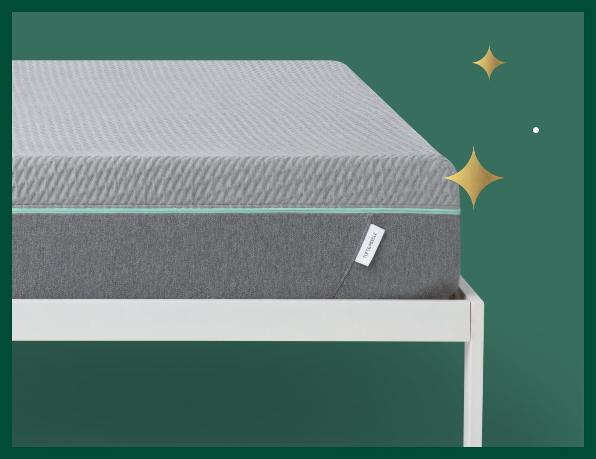 Corner of the Tuft & Needle Mint mattress sitting on a white bed frame in front of a green background with gold stars