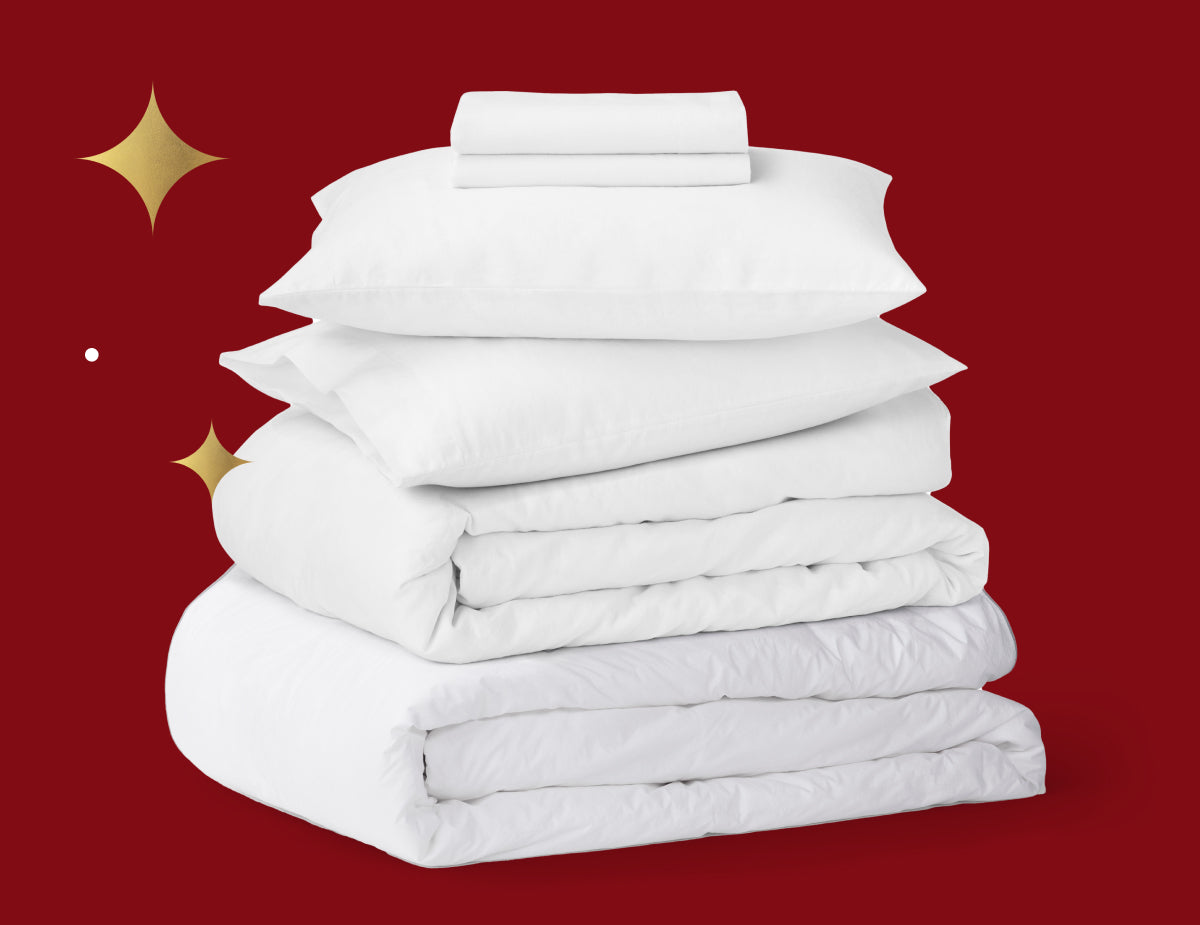 Tuft & Needle white linen duvet insert and cover folded, 2 white pillows, and linen sheets folded all stacked on top of each other. Picture has a red background with gold stars.