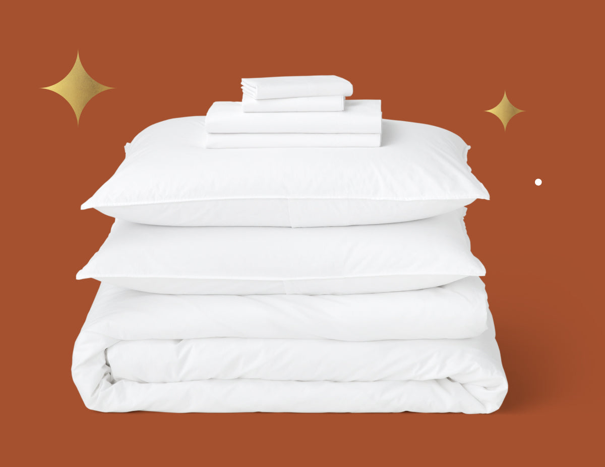 Tuft & Needle white duvet insert folded, 2 white pillows and a white percale sheet set folded all stacked on top of each other. Picture has a rust background with gold stars.