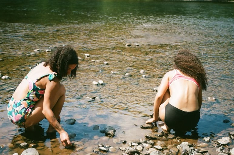 two people squatting at the shores of a stream, playing in the water.