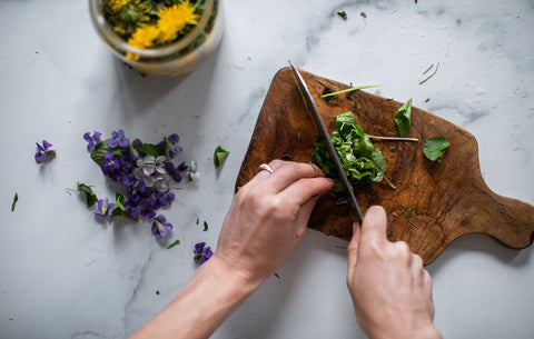 hands, holding a knife and chopping herbs atop a wooden cutting board. flowers and jars in the background.