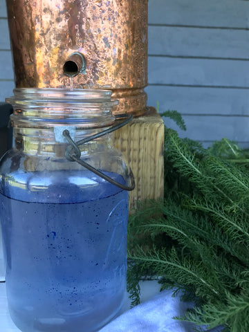 liquid flecked with blue essential oils collect in a jar at the base of a copper sprout