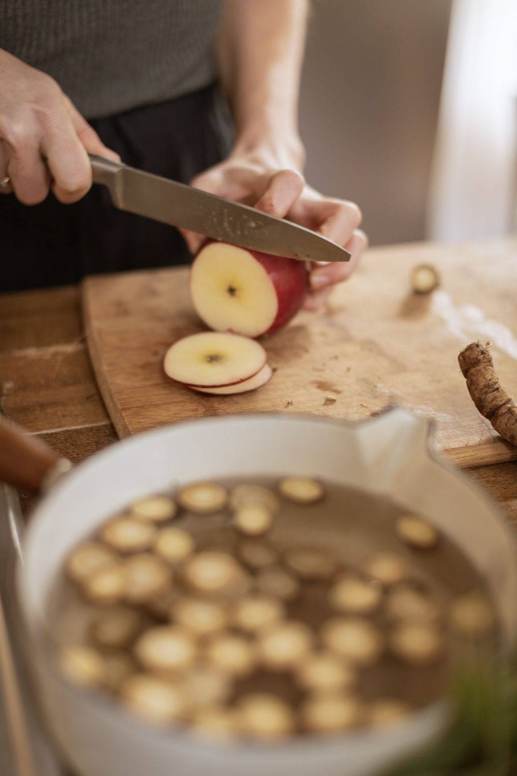 hands cutting an apple, a bot of tea with roots in it in the foreground.