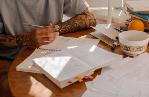 man with tattoos at a table covered in papers writing down a list