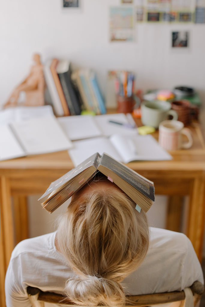 women with blonde hair at desk with head back and book on her face in defeat