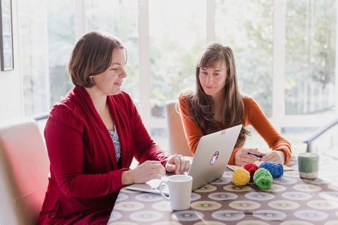 Helen Townsend and Anthea Madill sitting at a table looking at a laptop with EcoSplat Reusable Water Balloons on the table in front of them. Red balloon