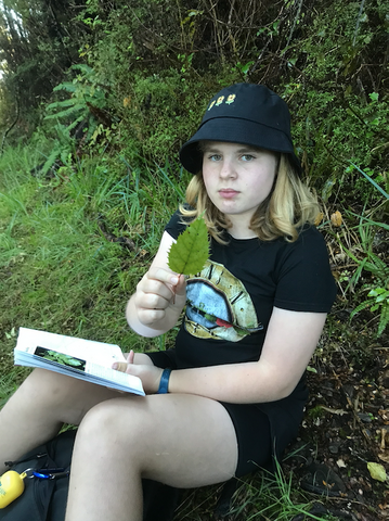 A 11 year old girl sitting in the forest holding a leaf up to the camera. She has a tree identification book in her lap and is wearing a black bucket hat and a Guns N' Roses t-shirt.