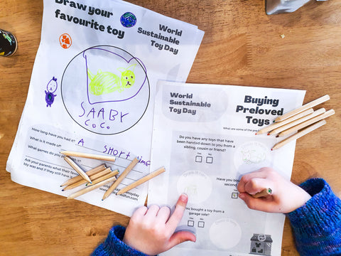 A bird's eye view of activity sheets on a table. There are coloured pencils and a girl's hands. The girl is holding a pencil and completing one of the sheets. The text on the sheets read "Draw your favourite toy" and "Buying pre-loved toys" and both sheets say "World Sustainable Toy Day".