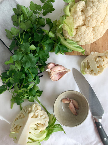Cauliflower, garlic and parsley spread out on a white tablecloth with linen grey napkins and a large knife.