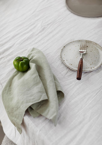 Sage green linen napkin on a white linen tablecloth alongside a plate, a wooden fork and green capsicum.