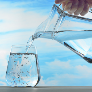 A hand pours water from a pitcher into a glass against a blue sky background.