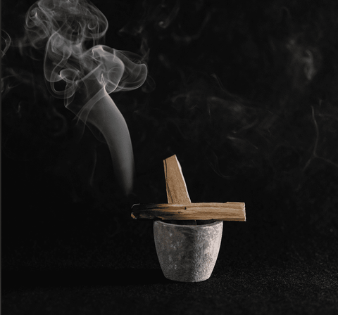 a stone bowl holding a smoldering piece of palo santo as smoke wisps through the air on a black backdrop