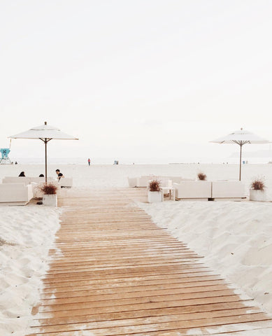 an image of a boardwalk stretching outward toward the water slowly encapsulated by sand. midway through the boardwalk are two sets of lounge couches accompanied by an umbrella