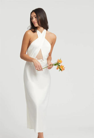 a woman stands on a white background. Her head is facing left away from the camera as she holds a bunch of flowers. She is in an ankle length, ivory night gown. The night gown has a halter neck, with a keyhole center, and on the waist is a floral applique.