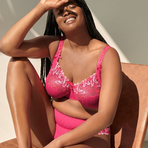 a Black woman, with long hair sits in a chair with her right elbow resting on her right leg. She is in a bright pink bra and panty set and is smiling