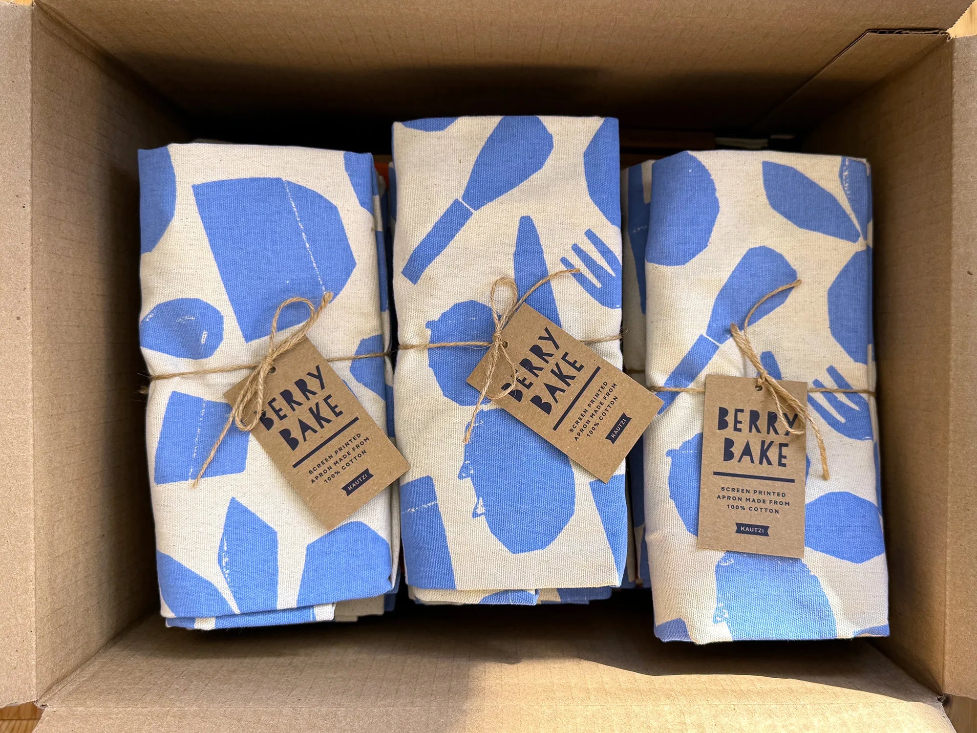 Three Berry Bake aprons, folded and neatly packed in a cardboard box. The aprons feature a blue and white baking-themed pattern and are tied with twine, each with a Kraft paper label attached.