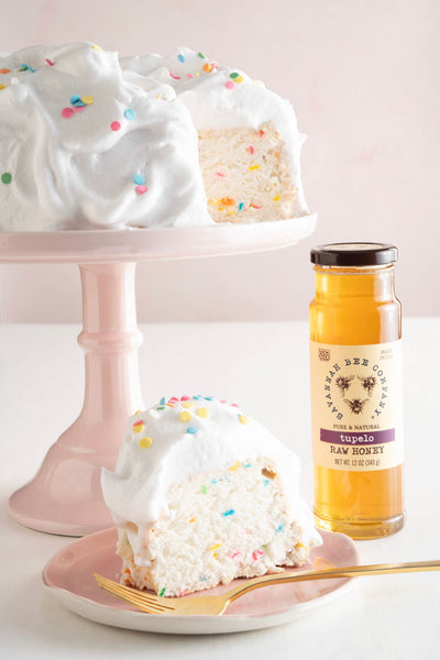 Angel cake with confetti frosting on a cake stand next to a 12 ounce jar of tupelo honey.