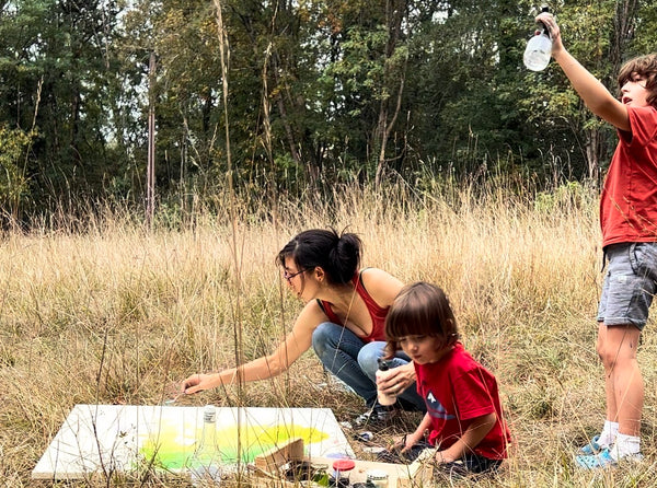 Une maman artiste dans un champ de la campagne française travaillant sur une peinture en techniques mixtes avec ses deux jeunes fils.