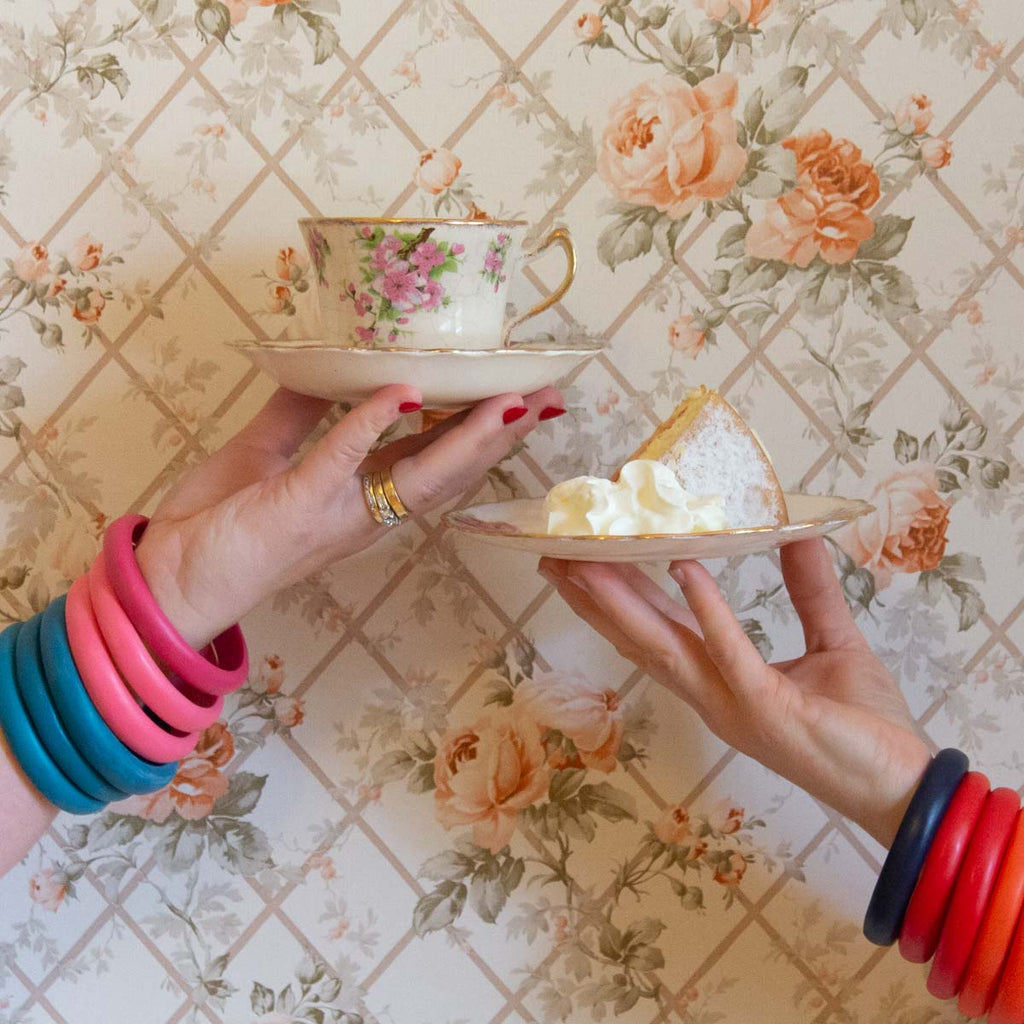 Hands holding a slice of cake and a cup of tea against a vintage rose wallpaper.