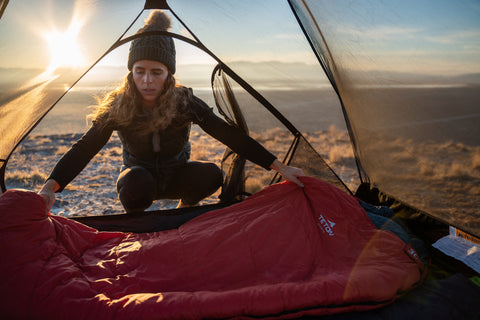 A woman spreads out a TETON Sports LEEF Sleeping Bag inside of her Mountain Ultra tent while the sun rises in the background.