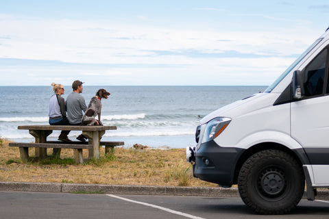 A couple and their dog sit beside the ocean on a picnic table with their cargo van in the foreground.