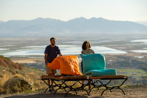 Two people unfurl their TETON Sports Celsius Sleeping Bags onto Outfitter XXL Camp Cots on a mountaintop.