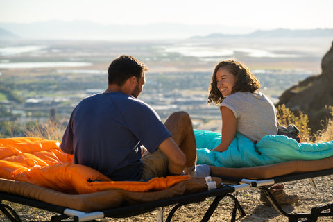 A couple laughs together while sitting atop their TETON Sports Celsius Sleeping Bags and Camp Cots.