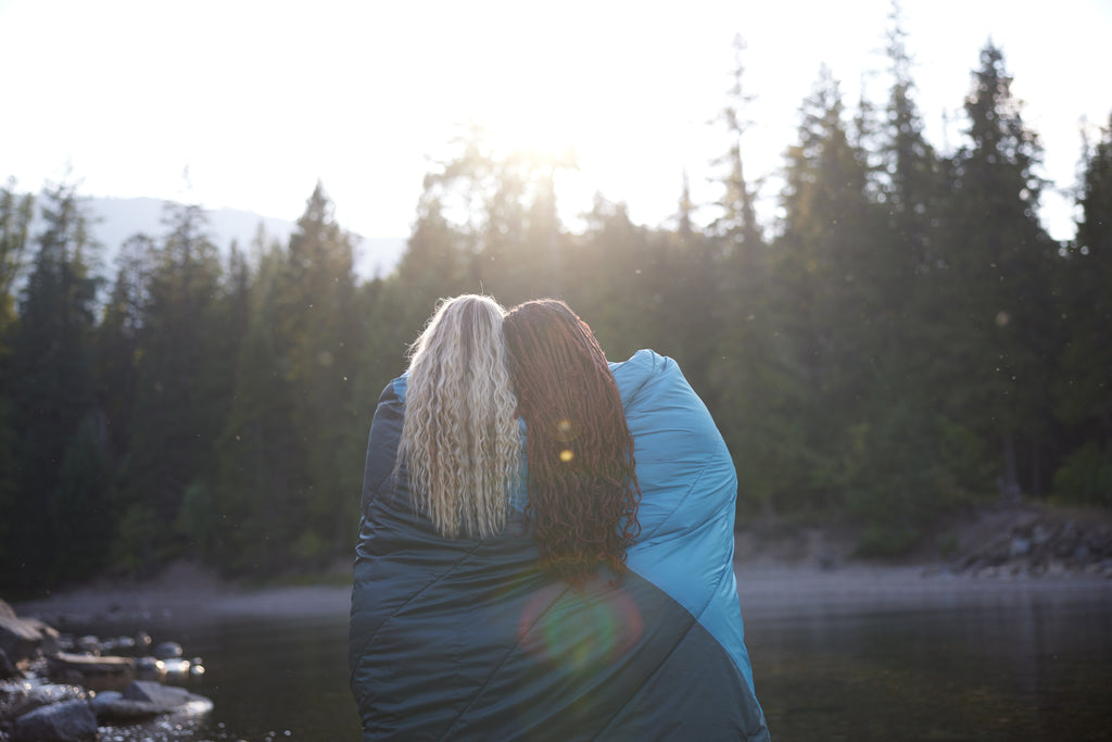 Two girls stand together in camp blanket looking at lake.
