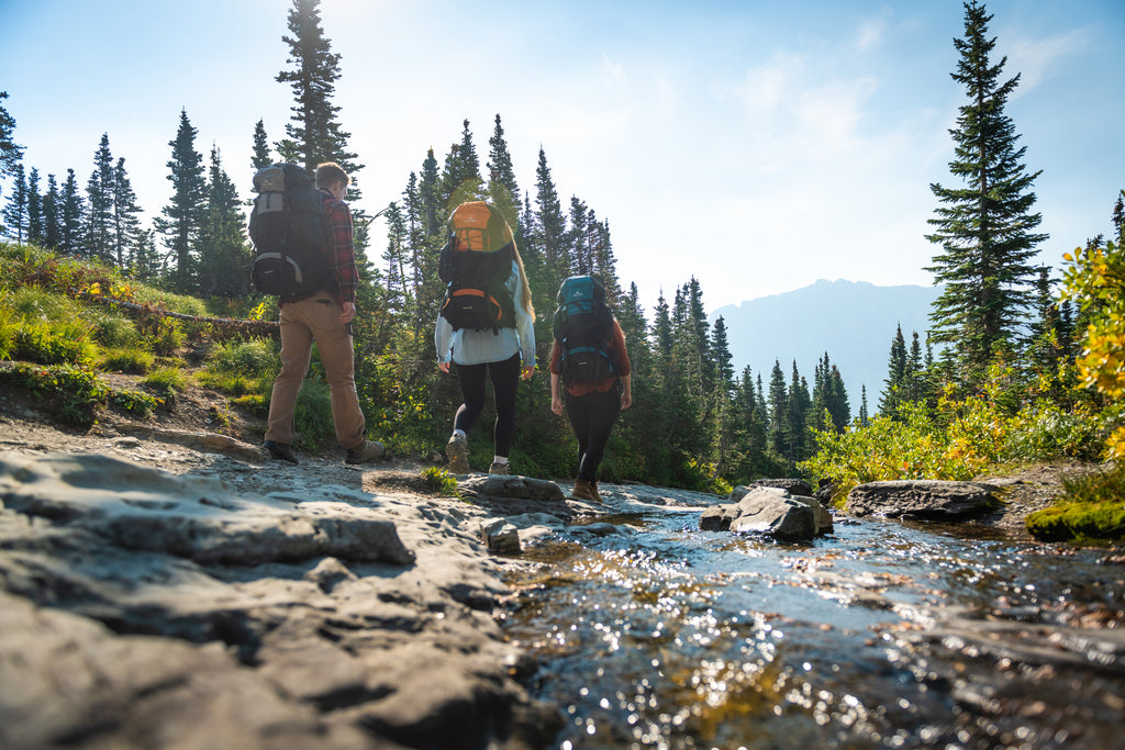 Three people walk near creek with backpacks. 