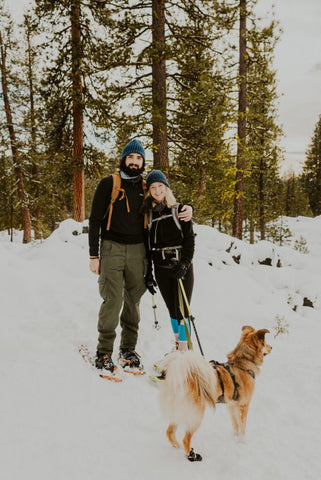 A man and a woman wearing snowshoes, posing while hold their dog on a leash in deep winter snow.