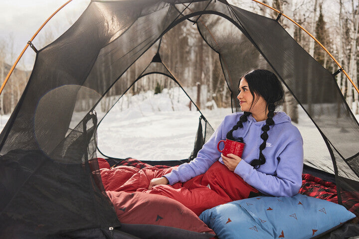 Woman sits in tent with mug in winter.