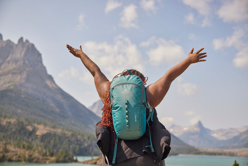 Woman stands on cliff with arms in the air with backpack.