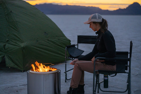 A woman roasts food over a campfire at twilight.