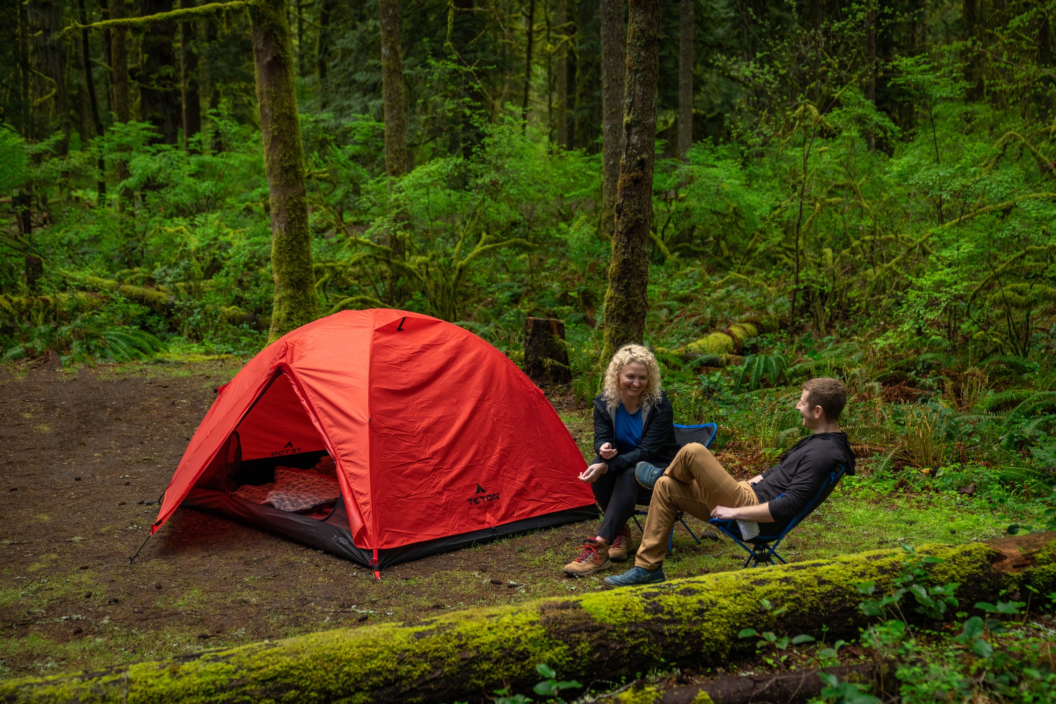 A couple sits together beside their TETON Sports Mountain Ultra Tent in red in a large forest.