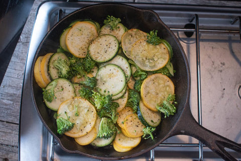 Zucchini and broccoli cooking over a camp stove from above in a cast iron pan.