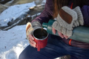 A gloved hand pours coffee from a Thermos into a camp mug.