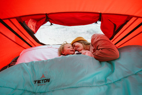 A mother and child snuggle together in a TETON Sports Cascade Two-Person Sleeping bag inside a tent.