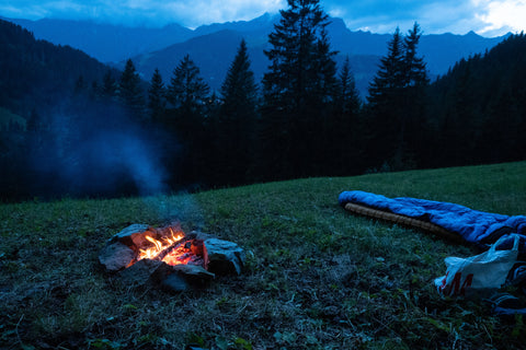 A campfire at twilight with an empty sleeping bag beside it.