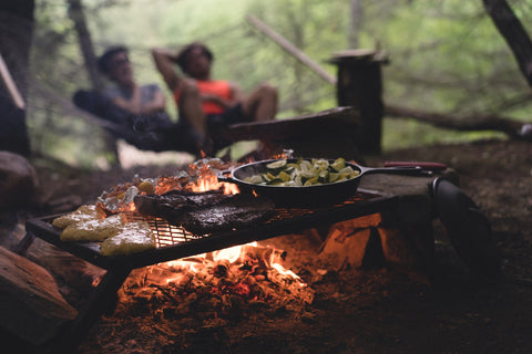Veggies cook in a pan on a campfire while a couple swings on a hammock together in the background.